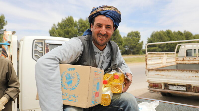 Man in headscarf sitting and smiling, holding a box of WFP food and bottles of oil